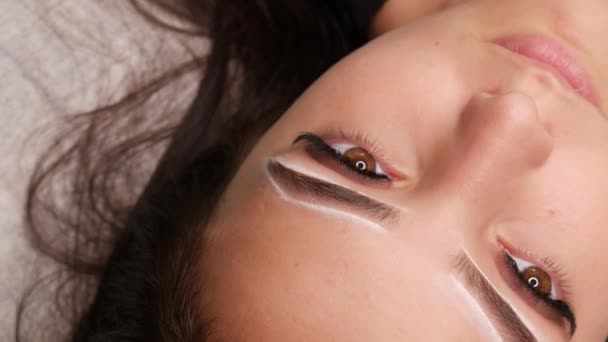 Master applique des marques blanches pour corriger la forme des sourcils avec une brosse spéciale. Le beau visage de la jeune fille modèle sur le canapé dans un salon de beauté avant microblading, tatouage permanent de sourcil de maquillage — Video