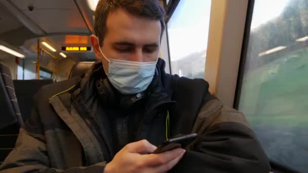 Joven guapo con una máscara médica, protección contra sarpullidos covid, monta en un tren y mira la pantalla de un teléfono inteligente — Vídeo de stock