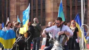 Strasbourg, Fransa - 26 Mart 2022: Rusya ile savaşa karşı Ukrayna 'yı destekleyen gösteri. Bir müzisyen Ukrayna bayraklarına karşı müzik aleti çalıyor ve posterler savaşı durduruyor..