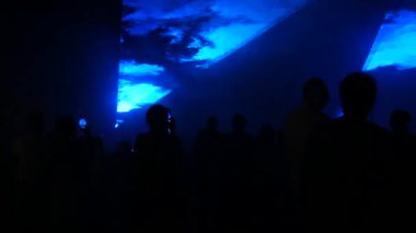 Modern bir lazer ışığı gösterisi, dumanla yansıtılan bir projeksiyonla, bir grup insan çeşitli çizgilere ve şekillere dönüşen lazer ışınlarını izliyor. Açık hava performansı