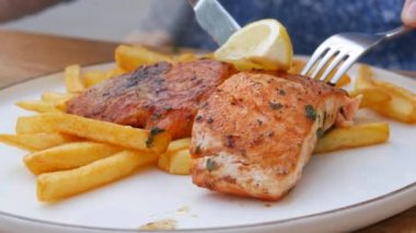 Büyük bir parça kırmızı somon balığı patates kızartması ve bir dilim limonun yanında ızgara yapılır. Bir kadın, beyaz bir tabakta çatal ve bıçakla bir parça kırmızı balığı açık bir kafe verandasında yer..