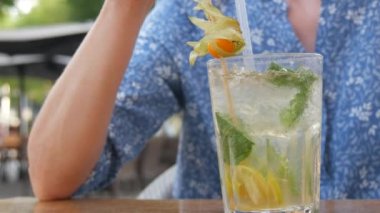 Yaz kokteyli. Şeffaf cam bardakta mojito kokteyli, içinde buz, nane ve limon yaprağı olan bir kadın, bir yaz kafesinin verandasındaki kokteyl tüpünden soğutucu bir kokteyl içer..