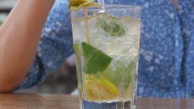 Yaz kokteyli. Şeffaf cam bardakta mojito kokteyli, içinde buz, nane ve limon yaprağı olan bir kadın, bir yaz kafesinin verandasındaki kokteyl tüpünden soğutucu bir kokteyl içer..