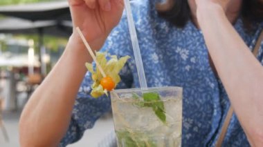 Yaz kokteyli. Şeffaf cam bardakta mojito kokteyli, içinde buz, nane ve limon yaprağı olan bir kadın, bir yaz kafesinin verandasındaki kokteyl tüpünden soğutucu bir kokteyl içer..