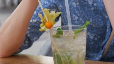 Yaz kokteyli. Şeffaf cam bardakta mojito kokteyli, içinde buz, nane ve limon yaprağı olan bir kadın, bir yaz kafesinin verandasındaki kokteyl tüpünden soğutucu bir kokteyl içer..