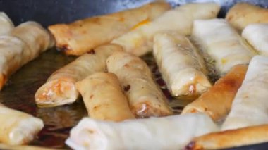 Asian spring rolls are fried in oil in a pan and turned over with kitchen tongs.