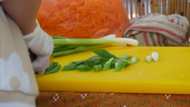 Female hands cut a stalk of onion greenery with a large knife on a plastic cutting board.