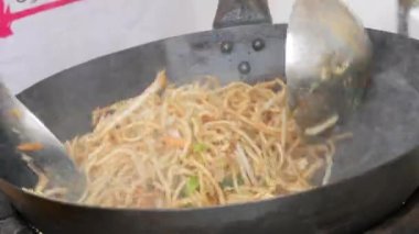 A female cooks hand frying a plate of thai noodles in a frying pan at an outdoor food festival.