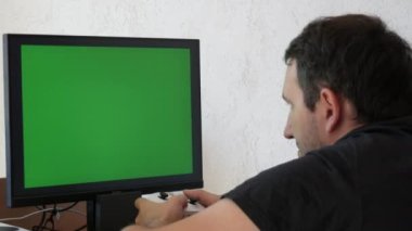Young male gamer playing game console using joystick remote control over green screen monitor tv chroma key at home. Man Focused Gaming in Living Room