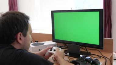 Young male gamer playing game console using joystick remote control over green screen monitor tv chroma key at home. Man Focused Gaming in Living Room