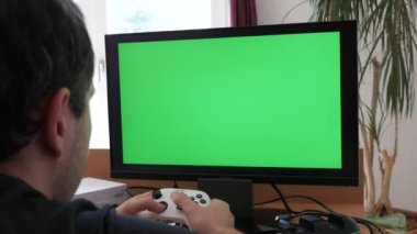 Young male gamer playing game console using joystick remote control over green screen monitor tv chroma key at home. Man Focused Gaming in Living Room