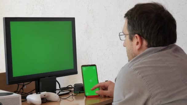 A young man in a shirt and glasses sits in front of a green chroma key monitor screen and clicks something on a smartphone with a green screen. Home office, freelancing in the living room at home