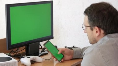 A young man in a shirt and glasses sits in front of a green chroma key monitor screen and clicks something on a smartphone with a green screen. Home office, freelancing in the living room at home