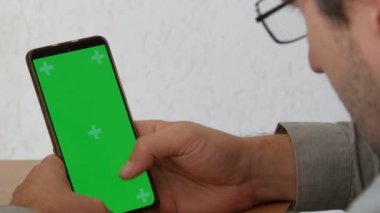 Green screen chroma key on a smartphone, the hands of a man in a shirt and glasses flipping through something on the screen.