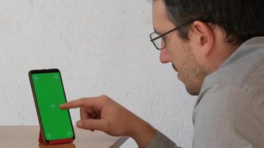 Green screen chroma key on a smartphone, the hands of a man in a shirt and glasses flipping through something on the screen.