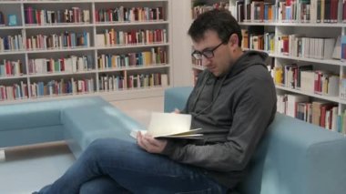 A young handsome intelligent man in glasses reads a book in a bookstore or library against the background of bookshelves. Student learning sitting on the steps