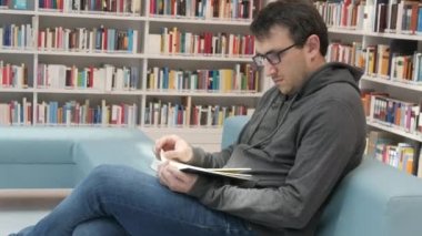A young handsome intelligent man in glasses reads a book in a bookstore or library against the background of bookshelves. Student learning sitting on the steps