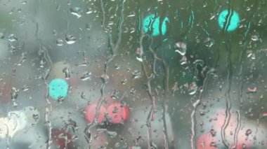 Rain on glass of a bus or car. Raindrops run down the window. Background of a city street, road. Drive car on traffic jam street at rain storm, inside a car driving,blurred traffic light background
