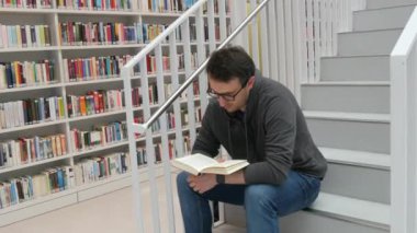 A young handsome intelligent man in glasses reads a book in a bookstore or library against the background of bookshelves. Student learning sitting on the steps