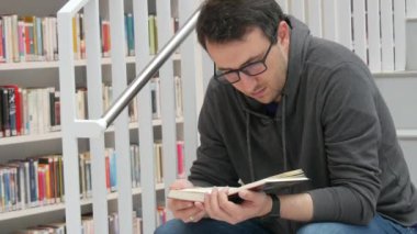 A young handsome intelligent man in glasses reads a book in a bookstore or library against the background of bookshelves. Student learning sitting on the steps