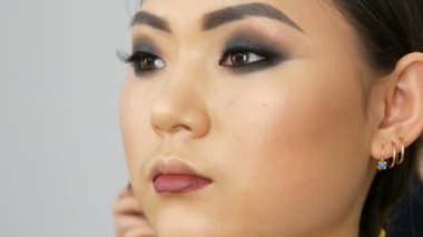 Professional stylist make up artist with special brush paints dark red lips with a special brush of an Asian girl model face in visage studio close up view.