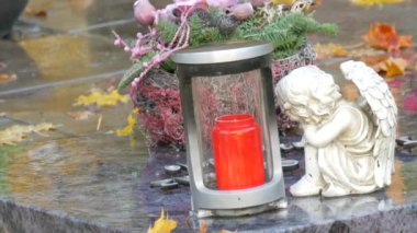 Red memorial candle burning with fire on rainy day on tombstone in cemetery autumn, rainy day. Grave with tombstone on raindrops falling, against abackground of yellow foliage naer sad angel statue.