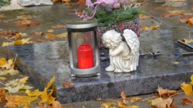 Red memorial candle burning with fire on rainy day on tombstone in cemetery autumn, rainy day. Grave with tombstone on raindrops falling, against abackground of yellow foliage naer sad angel statue.