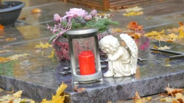 Red memorial candle burning with fire on rainy day on tombstone in cemetery autumn, rainy day. Grave with tombstone on raindrops falling, against abackground of yellow foliage naer sad angel statue.