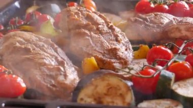 A large piece of chicken breast just from the oven baked with vegetables, cherry tomatoes, zucchini, eggplant, paprika. Steam comes from freshly cooked meat and vegetables on a baking sheet close up