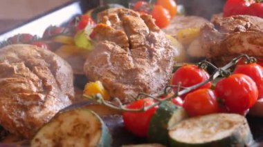 A large piece of chicken breast just from the oven baked with vegetables, cherry tomatoes, zucchini, eggplant, paprika. Steam comes from freshly cooked meat and vegetables on a baking sheet close up
