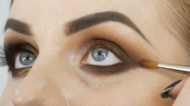 Makyaj ustası bir güzellik salonunda mavi gözlü model bir kadına profesyonel makyajlı altın dumanlı gözler uygular..