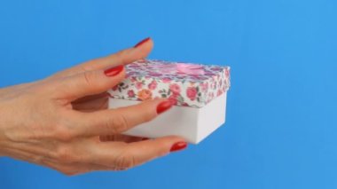 Bir çiçek baskı ve tırnakmavi bir arka plan üzerinde kırmızı oje ile boyanmış bir kadının elleri tarafından düzenlenen pembe bir yay ile küçük bir hediye kutusu.