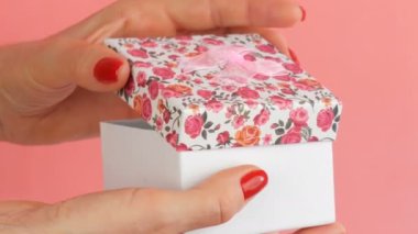 Bir çiçek baskı ve tırnakları pembe bir arka plan üzerinde kırmızı oje ile boyanmış bir kadının elleri tarafından düzenlenen pembe bir yay ile küçük bir hediye kutusu.