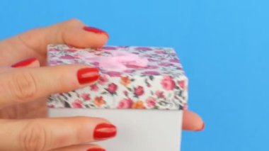 Bir çiçek baskı ve tırnakmavi bir arka plan üzerinde kırmızı oje ile boyanmış bir kadının elleri tarafından düzenlenen pembe bir yay ile küçük bir hediye kutusu.