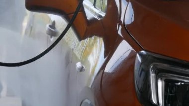 Sokakta turuncu bir arabanın yüzeyini özel bir su fırçası yıkıyor. Otomobil yıkama. Sabun köpüğü ve su damlası. Temizlik için özel fırça ve hortum.