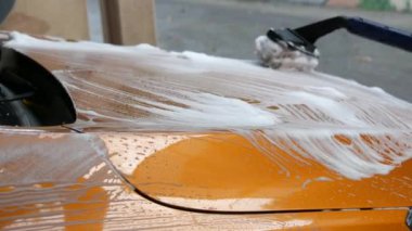 Oto yıkama. Sokakta turuncu bir arabanın yüzeyini özel sabunlu bir fırça yıkar. Otomobil yıkama. Sabun köpüğü ve su damlası. Temizlik için özel fırça ve hortum.