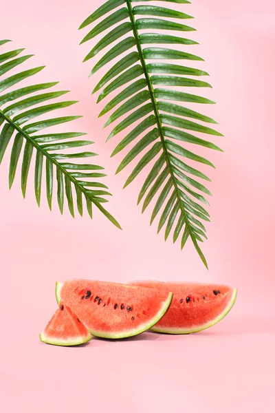 Creative Summer fruit party concept of refreshing watermelons under a palm tree. Pastel pink background.