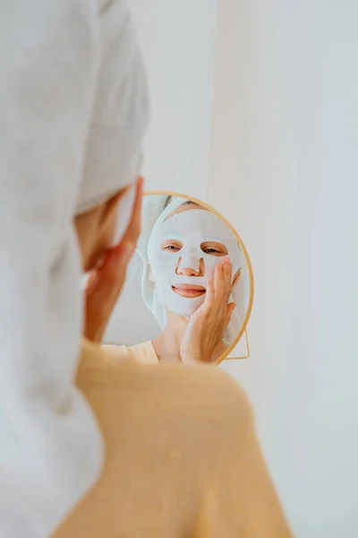 Woman with sheet moisturizing face mask looking in the mirror. Facial care and beauty treatments at home.