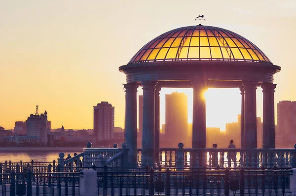 Rotunda of the city of Blagoveshchensk, Russia with a transparent dome and balustrade glows brightly in the rays of the setting sun. In the background, beyond the Amur River, the city of Heihe, China.