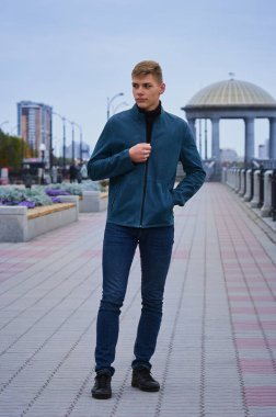 Şehirde gezinen genç bir adam bir sonbahar akşamında ceketinin düğmelerini ilikliyor. Arka planda bulanık binalar ve kubbeler var. Banklar ve set çitleri. Tam boy. Seçici odak.