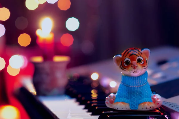 Uma estatueta de um filhote de tigre de brinquedo em uma camisola azul se senta nas teclas de um instrumento musical. Uma vela está a arder num castiçal. O brilho da guirlanda de Ano Novo. Decoração de árvore de Natal. Imagens De Bancos De Imagens Sem Royalties