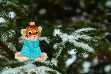 Mavi kazaklı bir kaplan yavrusu heykelciği dışarıdaki bir Noel ağacının karla kaplı dalında oturur. İğnelerde kar taneleri. Kaplan, 2022 yılının bir sembolüdür. Seçici odak.