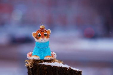 Mavi kazaklı kaplan yavrusu kışın kütüğün üstünde. Noel ağacı için oyuncak. Kar yağıyor. Şehir caddesi, ulaşım aracı çok bulanık. Tiger, Çin takviminde 2022 'nin sembolüdür. Seçici odak.