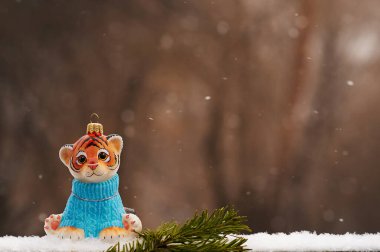 Kırmızı kaplan yavrusu, mavi süveter giymiş, üzerinde Noel ağacı dalı olan. 2022 yılının kaplan sembolü. Arka planda bulanık kahverengi orman. Kar yağıyor.
