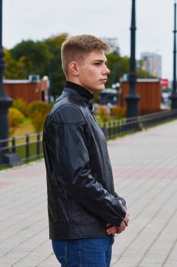 Dışarıda poz veren siyah ceketli genç adam. Arka planda sonbahar yapraklı ağaçlar bulanık.