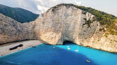Yunanistan 'ın Zakynthos kentindeki İyon Denizi kıyısındaki Navagio plajının insansız hava aracı görüntüsü. Kayalık kayalıklar, demirli kayıklar ve dinlenen insanlar, mavi su