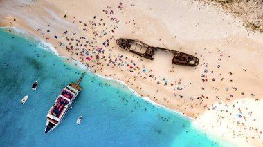 Yunanistan 'ın Zakynthos kentindeki İyon Denizi kıyısındaki Navagio plajının insansız hava aracı görüntüsü. Demir atmış tekneler, dinlenen insanlar, paslı gemi, mavi su.