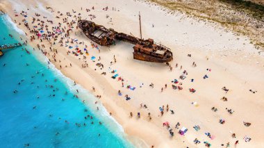 Yunanistan 'ın Zakynthos kentindeki İyon Denizi kıyısındaki Navagio plajının insansız hava aracı görüntüsü. Paslı bir geminin yanında dinlenen insanlar, mavi su.