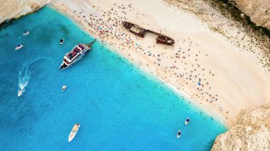 Yunanistan 'ın Zakynthos kentindeki İyon Denizi kıyısındaki Navagio plajının insansız hava aracı görüntüsü. Demir atmış tekneler, dinlenen insanlar, paslı gemi, mavi su.