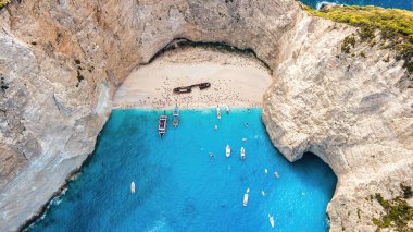 Yunanistan 'ın Zakynthos kentindeki İyon Denizi kıyısındaki Navagio plajının insansız hava aracı görüntüsü. Kayalık kayalıklar, demirli kayıklar ve dinlenen insanlar, mavi su